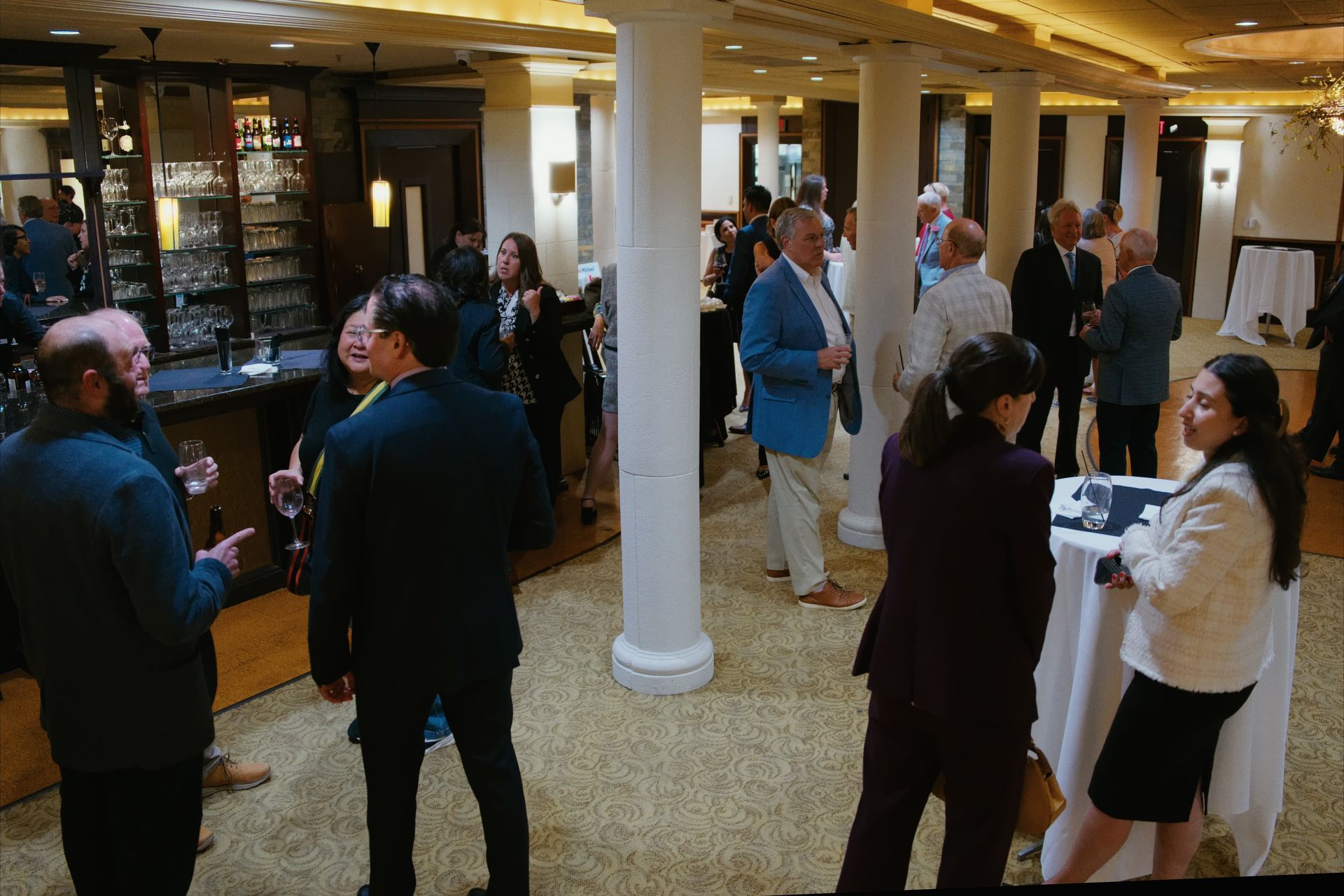 People mingling at a formal indoor reception with white columns and a bar in the background