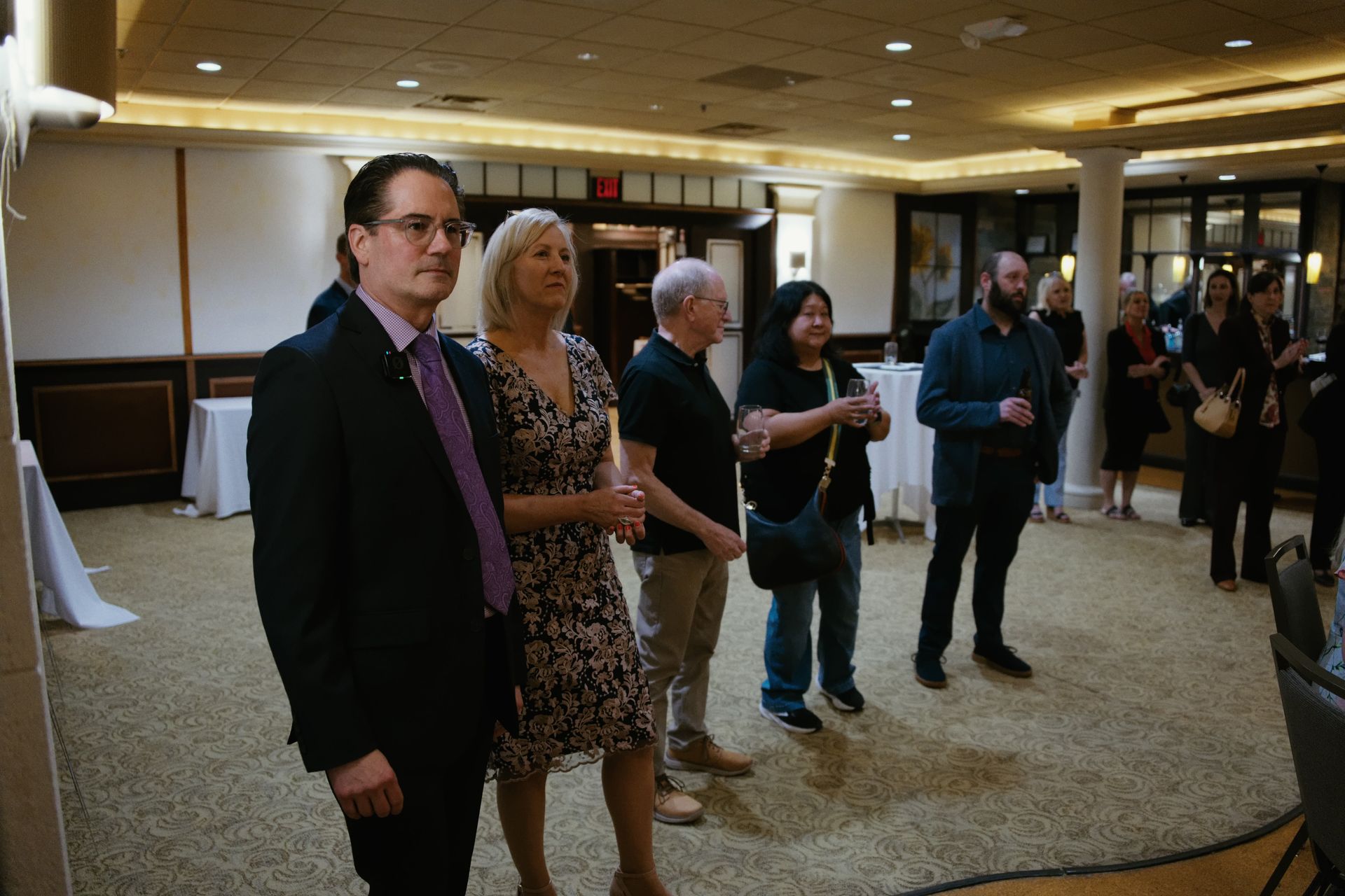 People standing in a hotel lobby, waiting and conversing near a carpeted reception area.