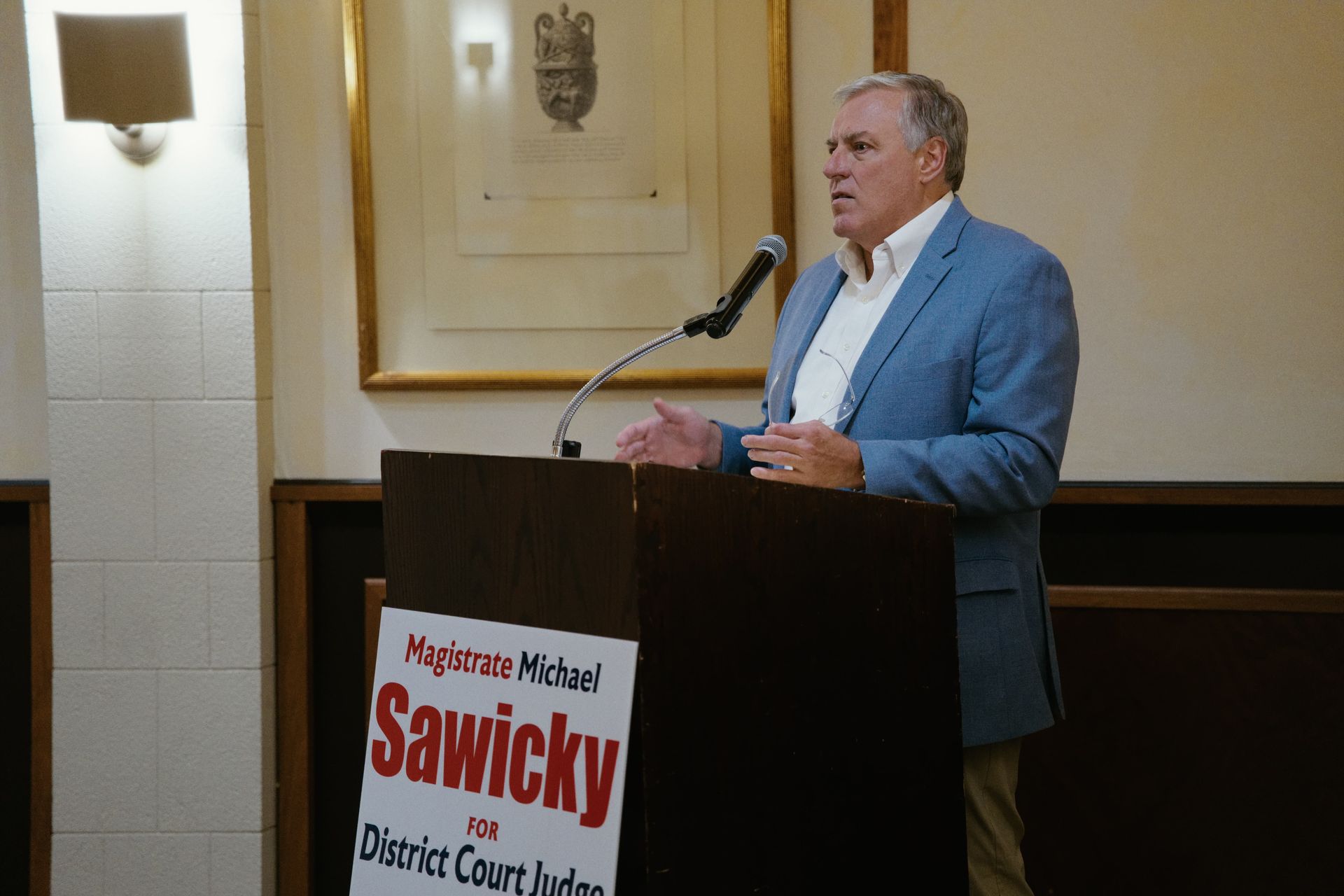 Man speaking at a podium with a campaign sign in a formal indoor room