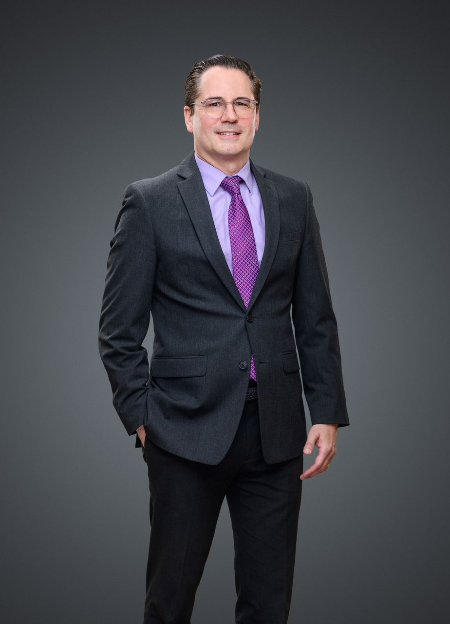 Sawicky in suit, smiling, standing with hand in pocket, against a gray backdrop.