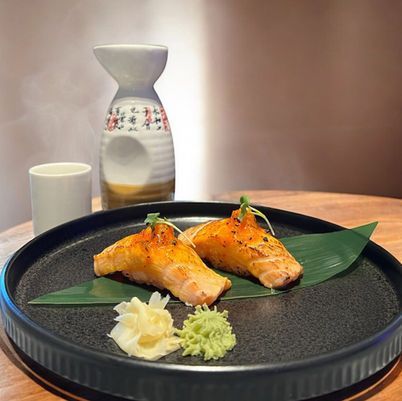 Plated grilled salmon on a black dish with green leaves and garnish, beside a cup and ceramic sake bottle.