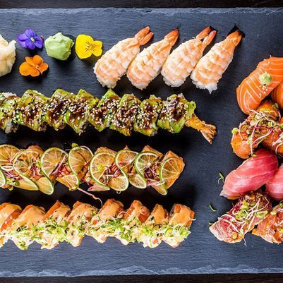 Assorted sushi rolls and nigiri on a black slate platter, with colorful garnishes on a dark table.