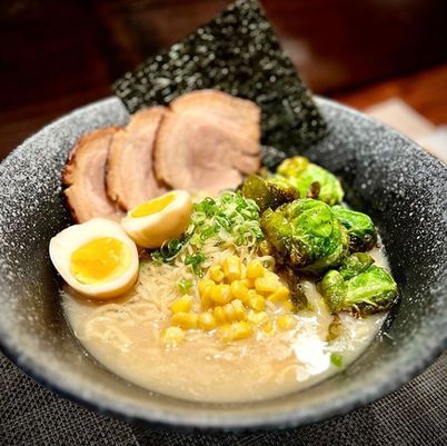 Ramen bowl with sliced pork, soft-boiled egg, corn, and Brussels sprouts on a table