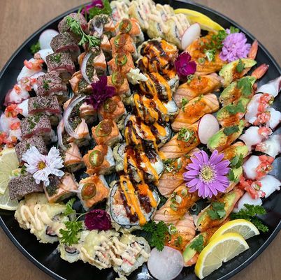 Assorted sushi rolls and sashimi on a black platter, garnished with flowers, lemon, and sauces