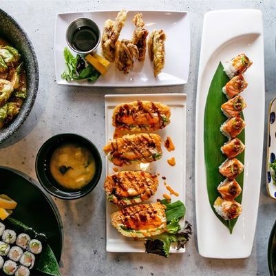 Assorted sushi and grilled dishes arranged on white plates with dipping sauces on a gray table.