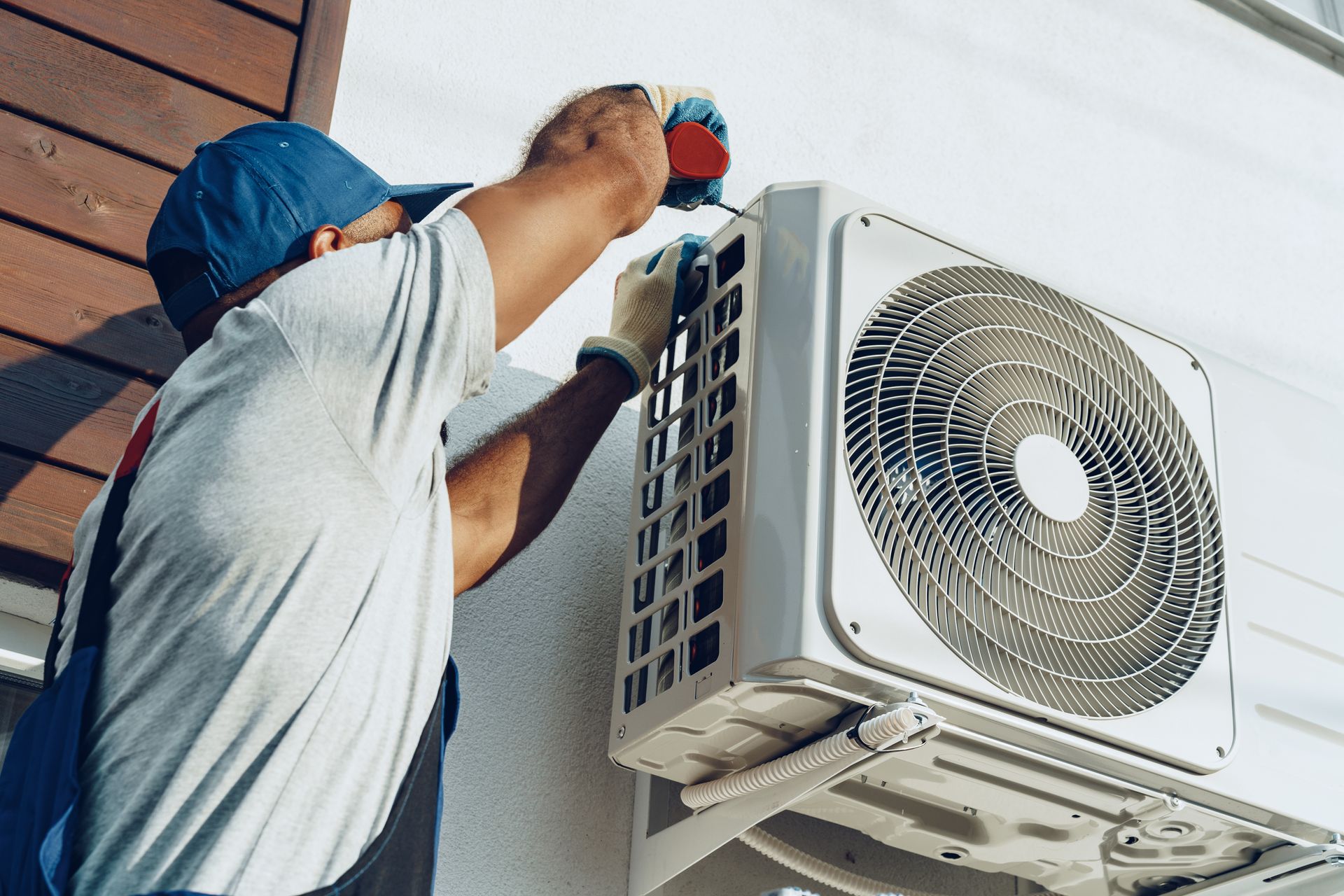 A technician installs an outdoor air conditioning unit on an exterior wall using power tools.