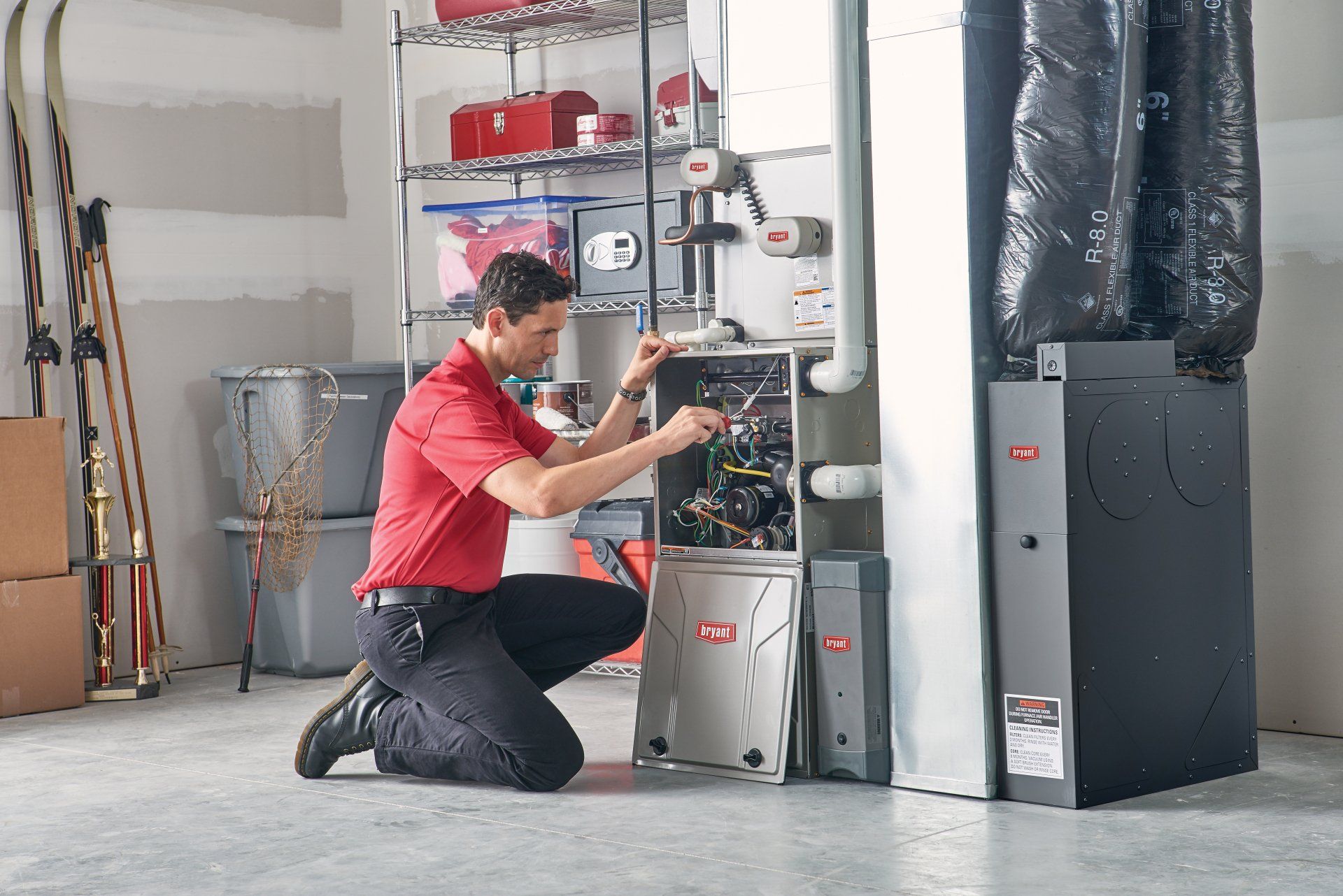 HVAC technician kneels, working on a furnace in a garage. Wearing red shirt, black pants.