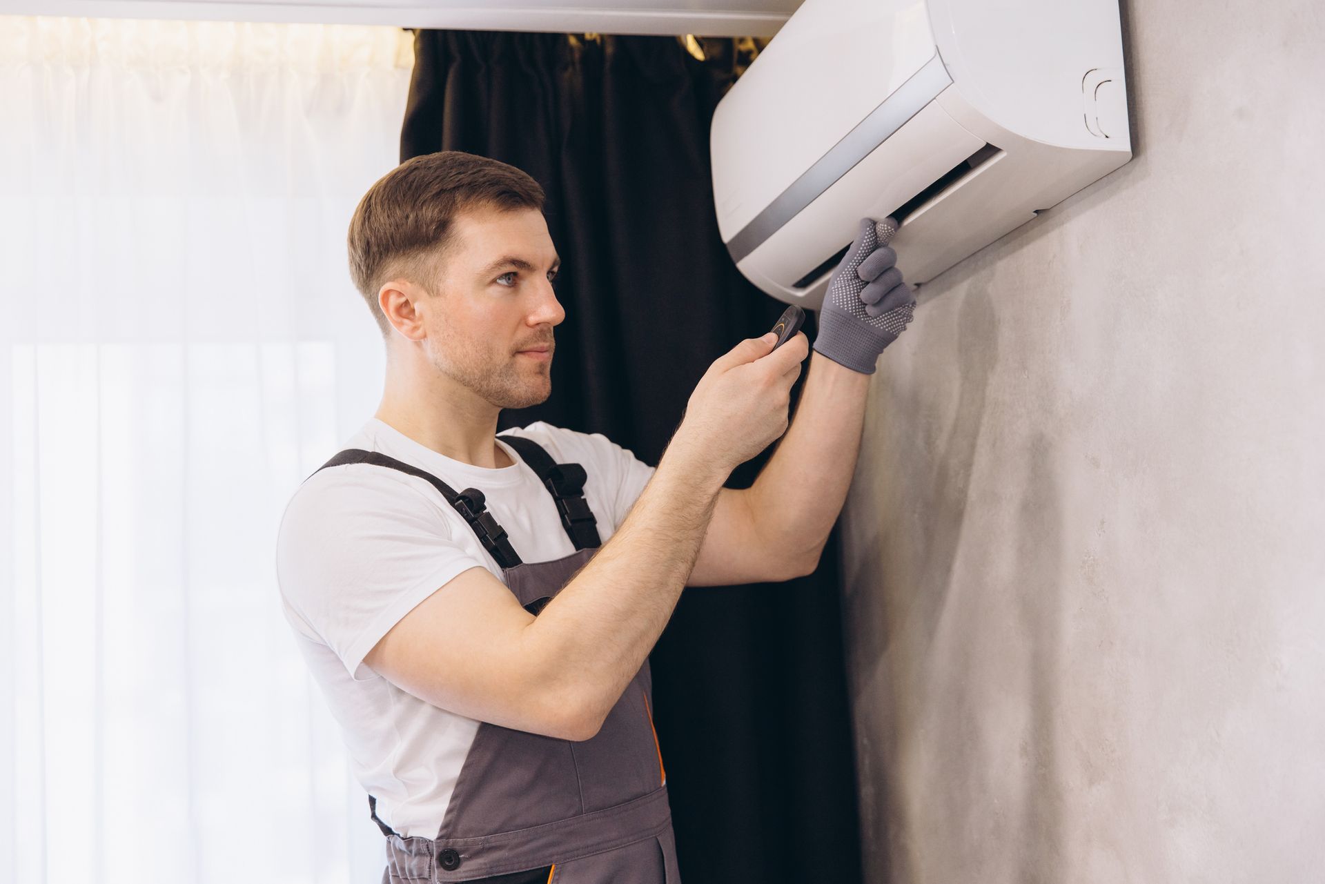 A technician in overalls repairing a wall-mounted air conditioner indoors