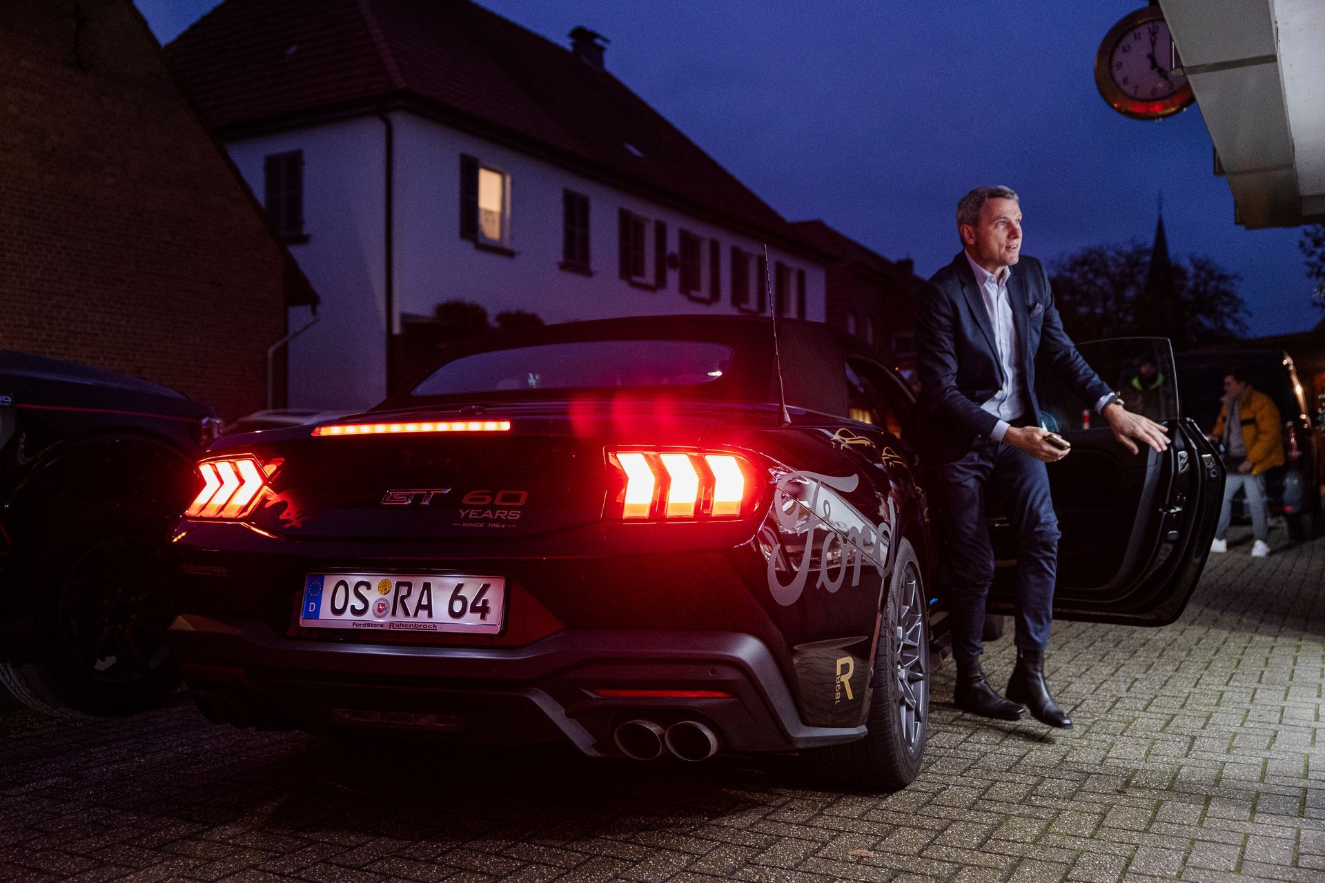 Ein Mann im Anzug steigt aus einem schwarzen Mustang aus, mit dem Nummernschild „OS-RA 64“