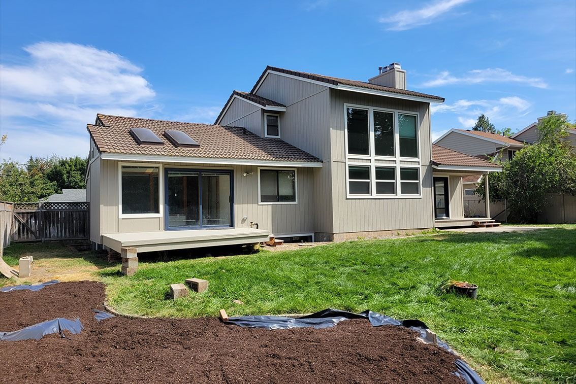 Backyard view of a damaged house with a large windowed extension and torn-up lawn work in the foreground