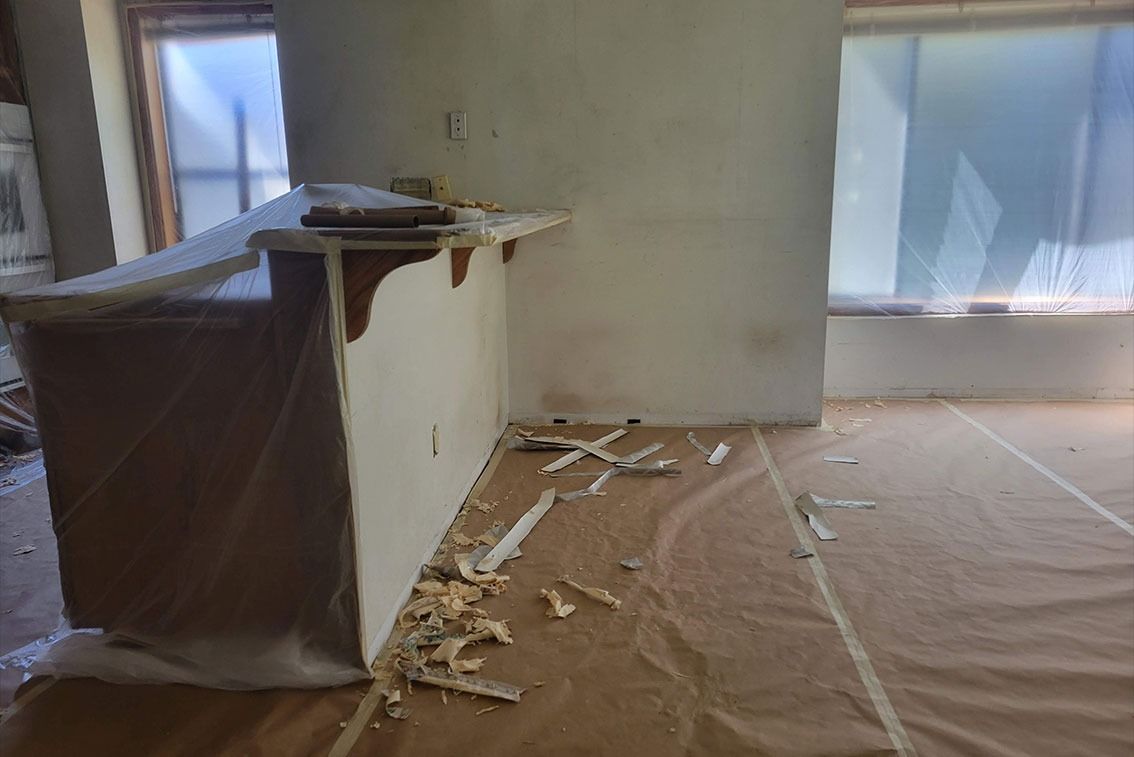 Damaged room with torn drywall, debris on the floor, and a covered cabinet near a window.