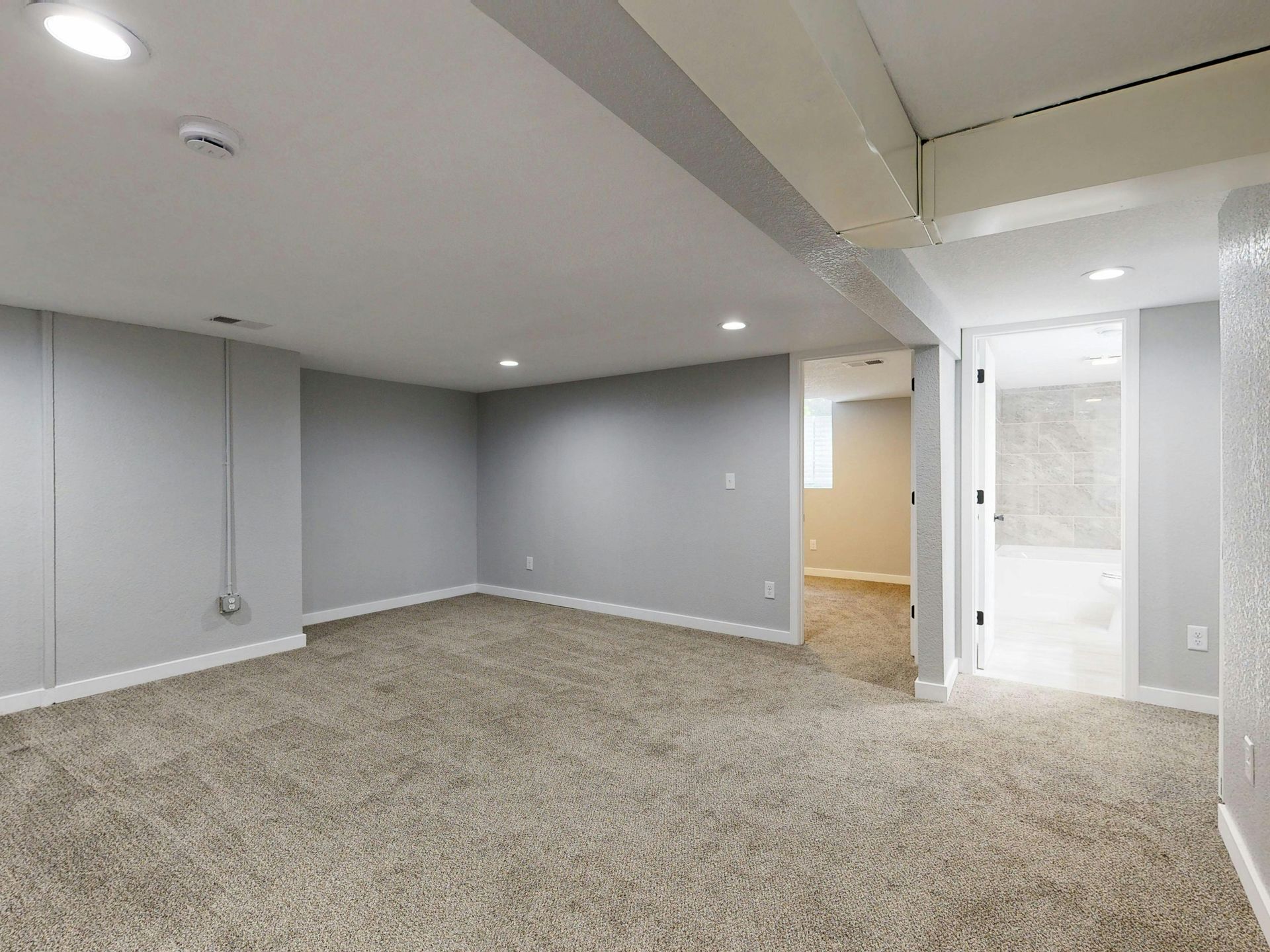 Empty carpeted basement room with gray walls, recessed lights, and a doorway to a hall