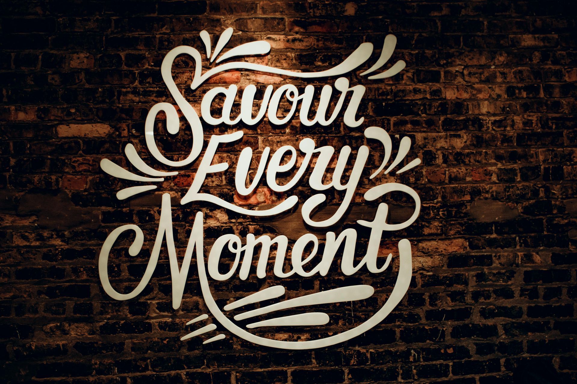Hand-lettered “Savour Every Moment” in white on a dark brick wall with leafy accents