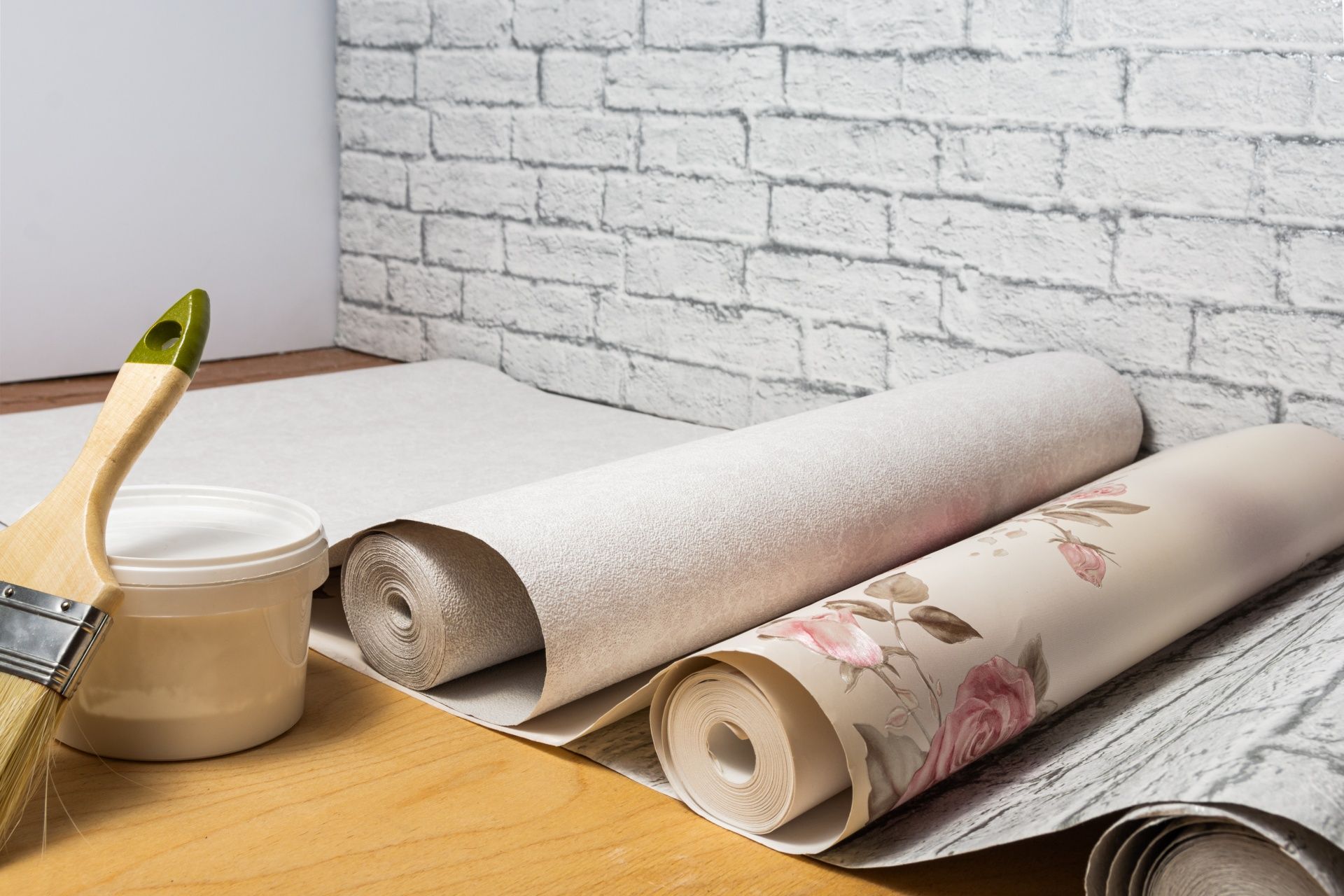 Paintbrush, wallpaper rolls, and paste on a wooden table against a white brick wall.