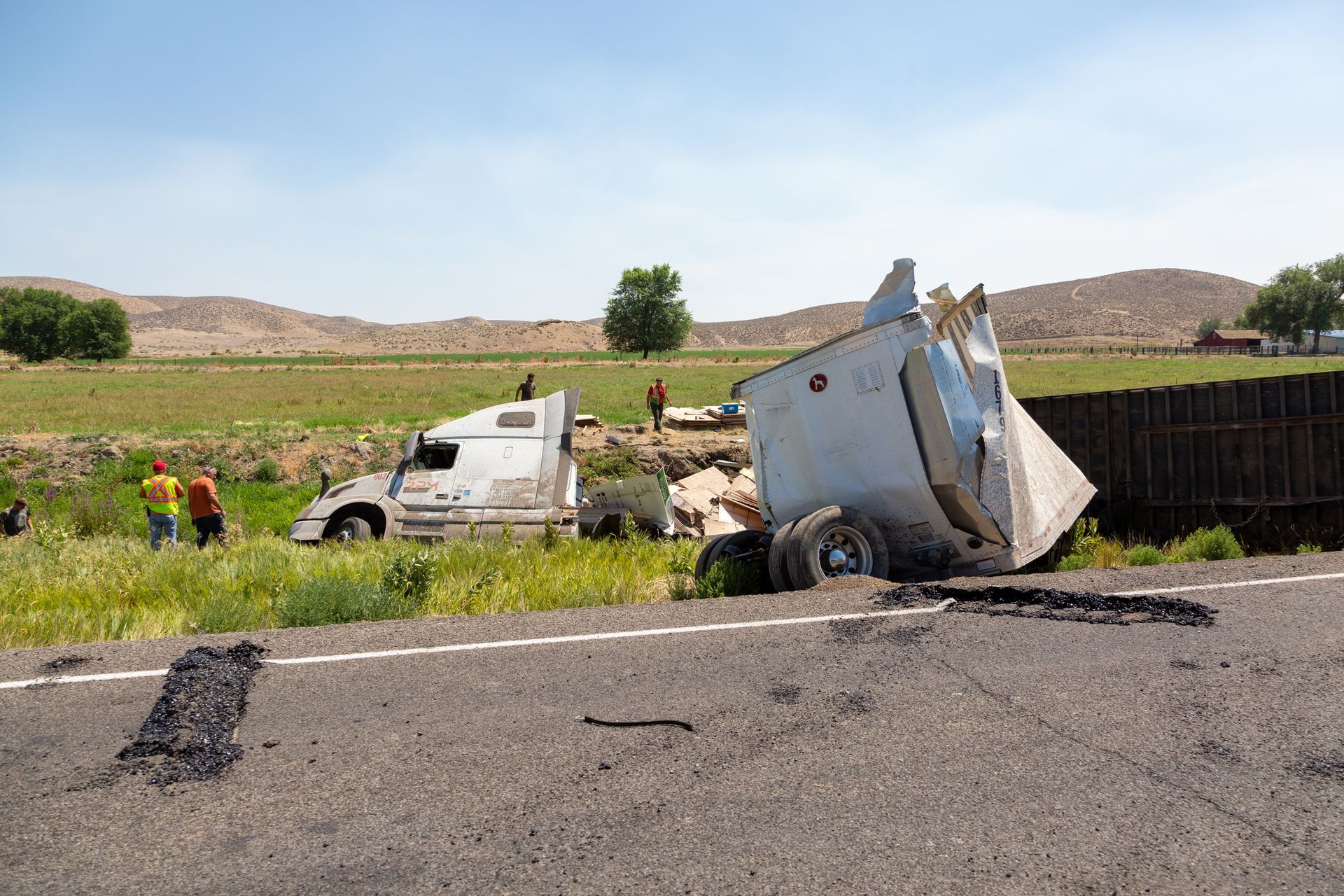 Semi-truck crashed off the road; white cab, damaged trailer. Workers assess the scene, grassy ditch, sunny day.