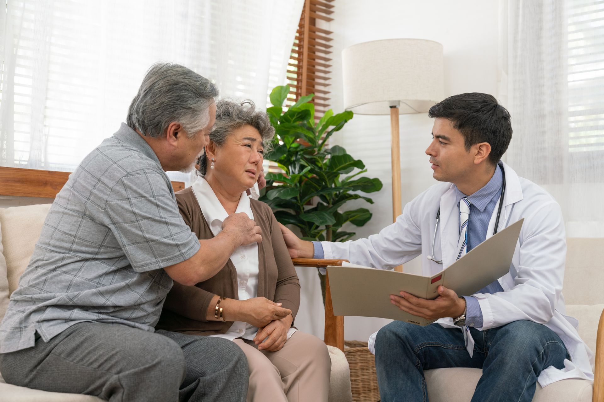 Doctor comforts elderly couple in a living room; the woman appears distressed, while the man offers support.