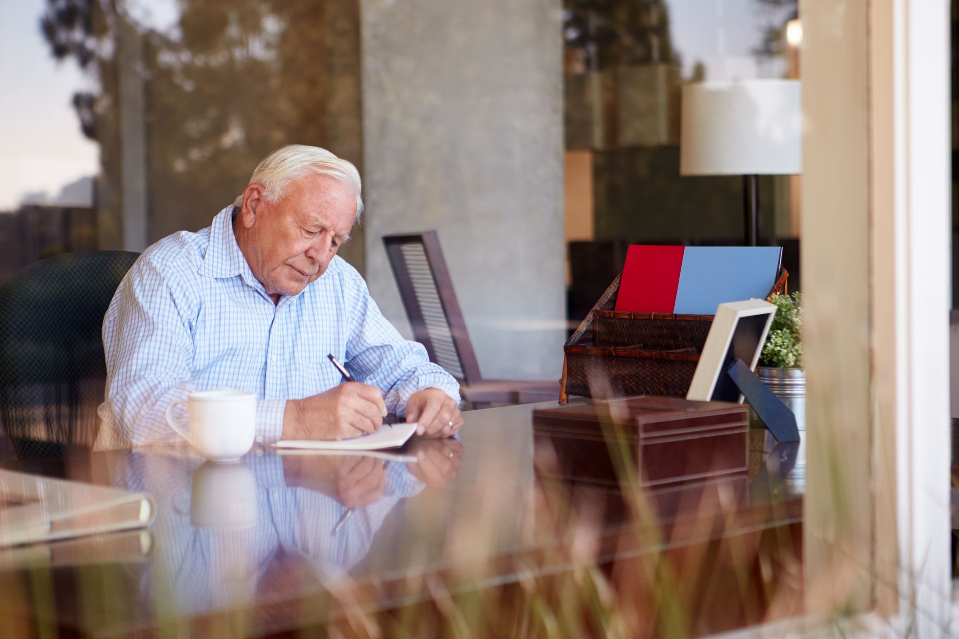Older man writing at a desk indoors, seen through a window. He wears a blue shirt, has a coffee cup and documents nearby.