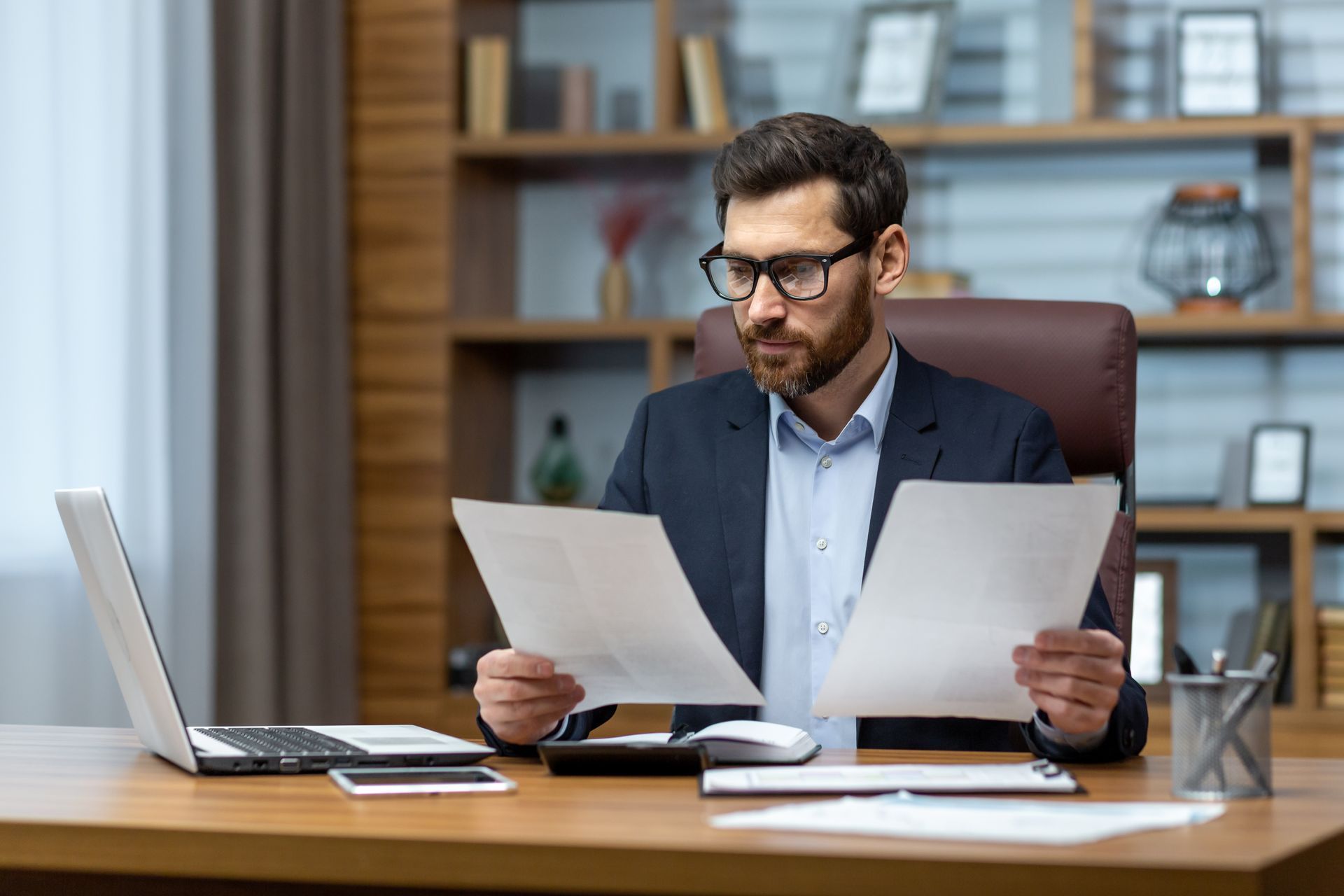 Man in suit at desk, reviewing papers, laptop, glasses, focused expression.