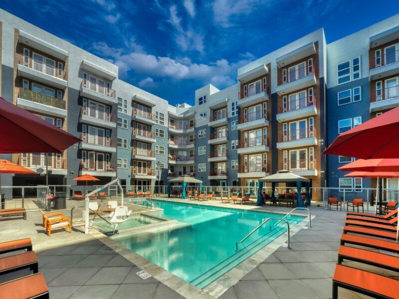 A large swimming pool surrounded by chairs and umbrellas in front of a large apartment building.