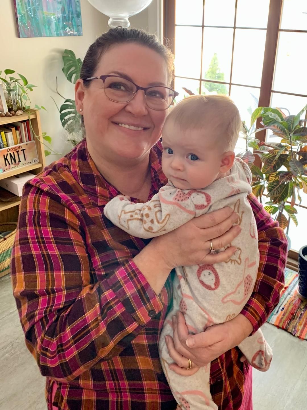 Woman smiling and holding a baby indoors near a bright window and plants