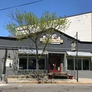 Small storefront with a white facade, red door, awnings, and a tree in front on a sunny day.