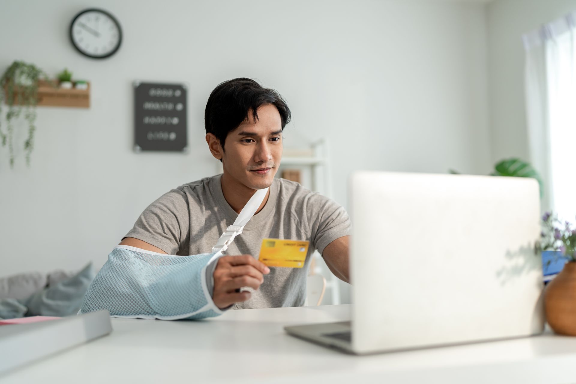 Man with arm in sling uses laptop, holding credit card. Indoors, white walls, clock.
