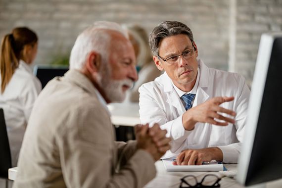 A healthcare professional in a white coat explains information on a computer screen to a patient in an office setting.