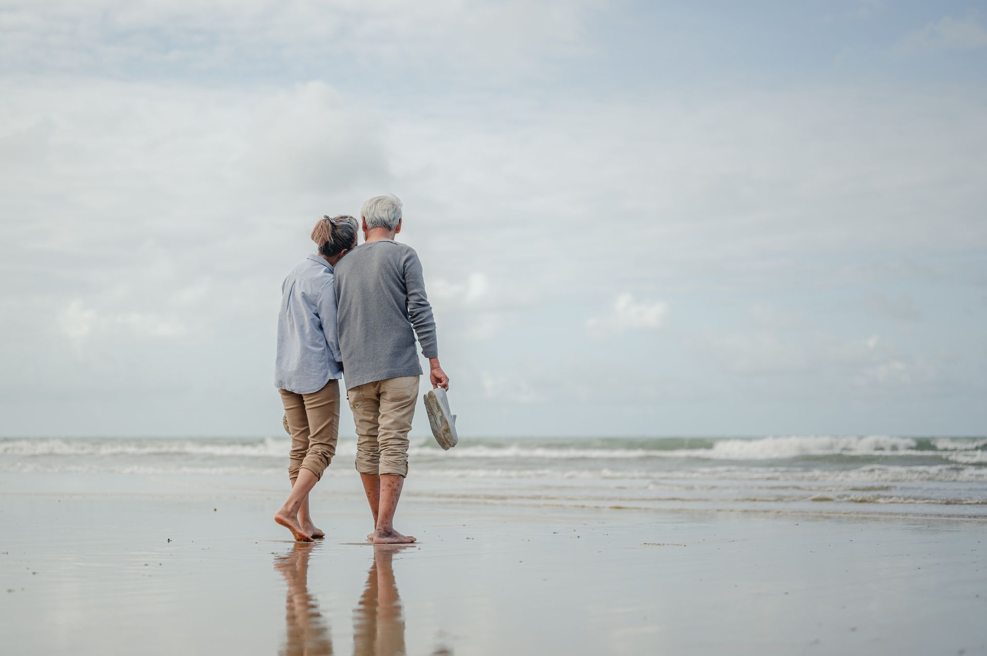 Two people walk hand-in-hand along a sandy beach, their reflections visible in the wet sand under an overcast sky.