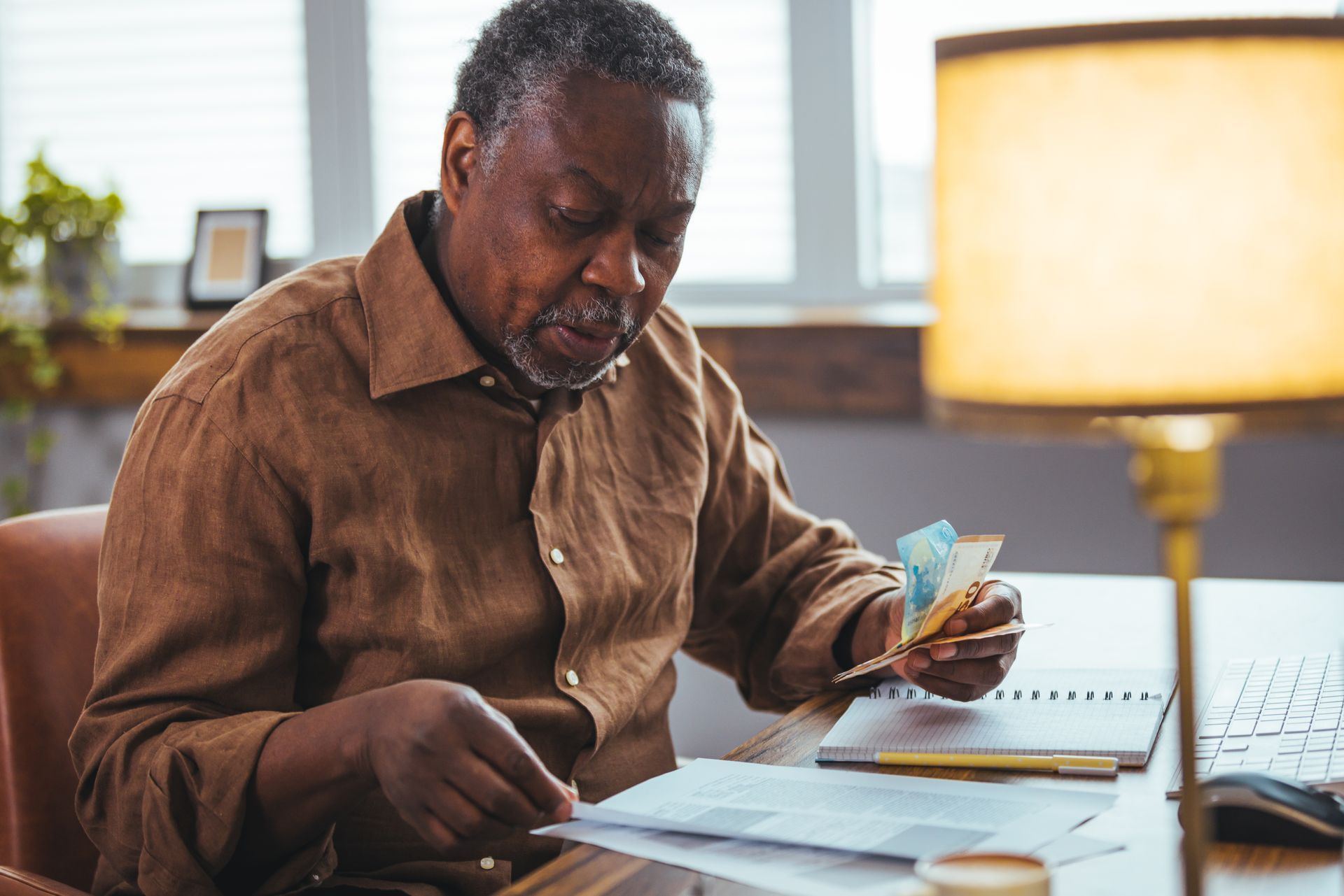 A person in a brown shirt sitting at a desk with papers and money, reviewing financial documents near a lamp.
