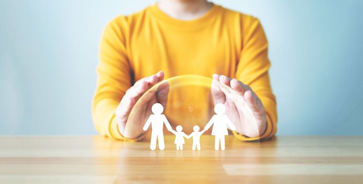 A person in a yellow sweater cups their hands over a paper cutout of a family, symbolizing protection and care.