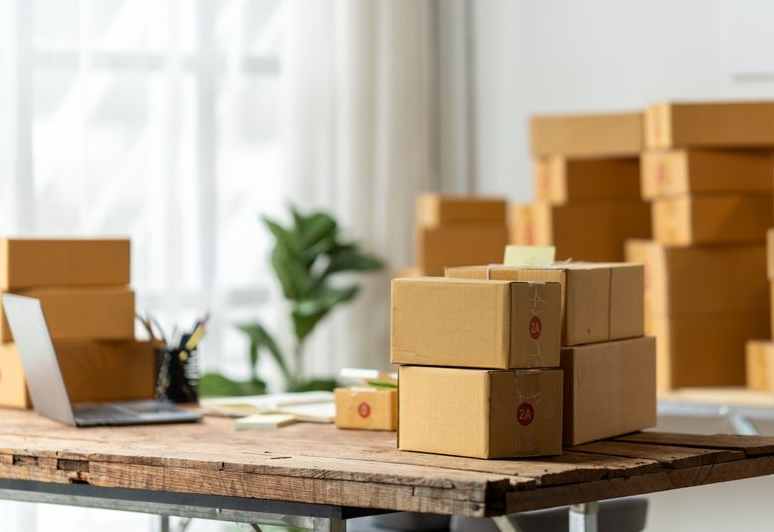 Packed cardboard boxes on a table in a bright room, with more boxes stacked in the background.