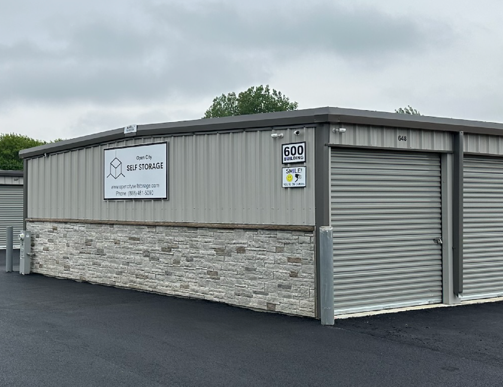 Gray storage building with roll-up doors and a sign, set on a paved lot under cloudy sky