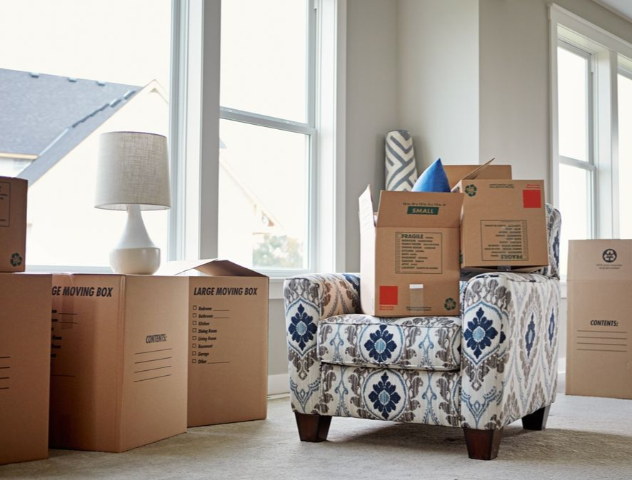 Living room packed with moving boxes around a patterned armchair near bright windows