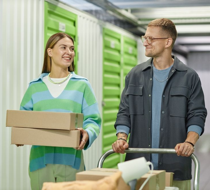Two people in a warehouse smiling while carrying boxes and pushing a cart