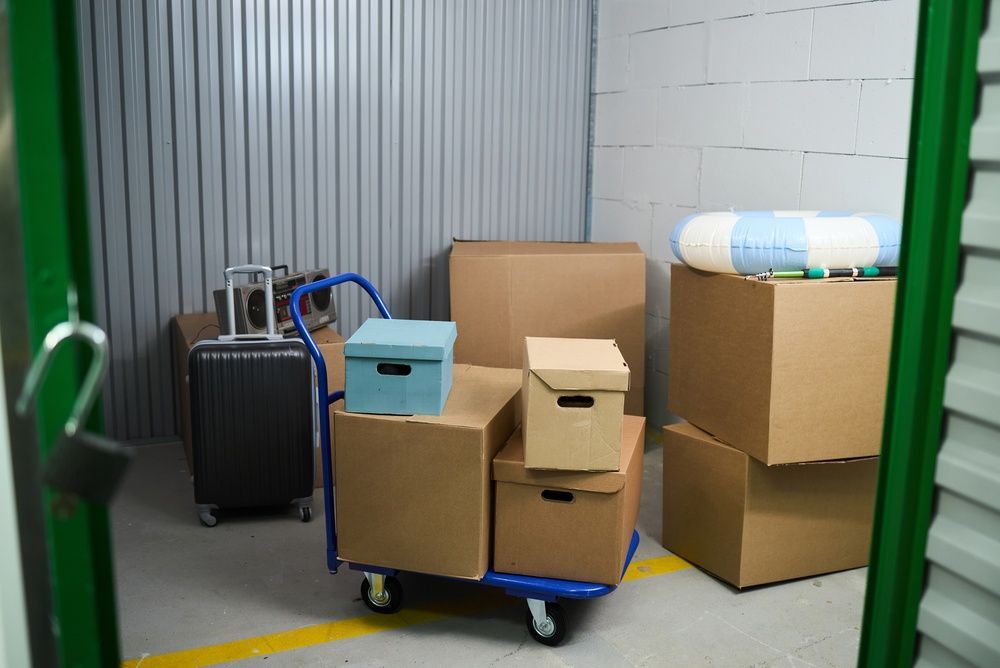 Boxes and suitcases stacked on a cart in a storage area with green shelving.