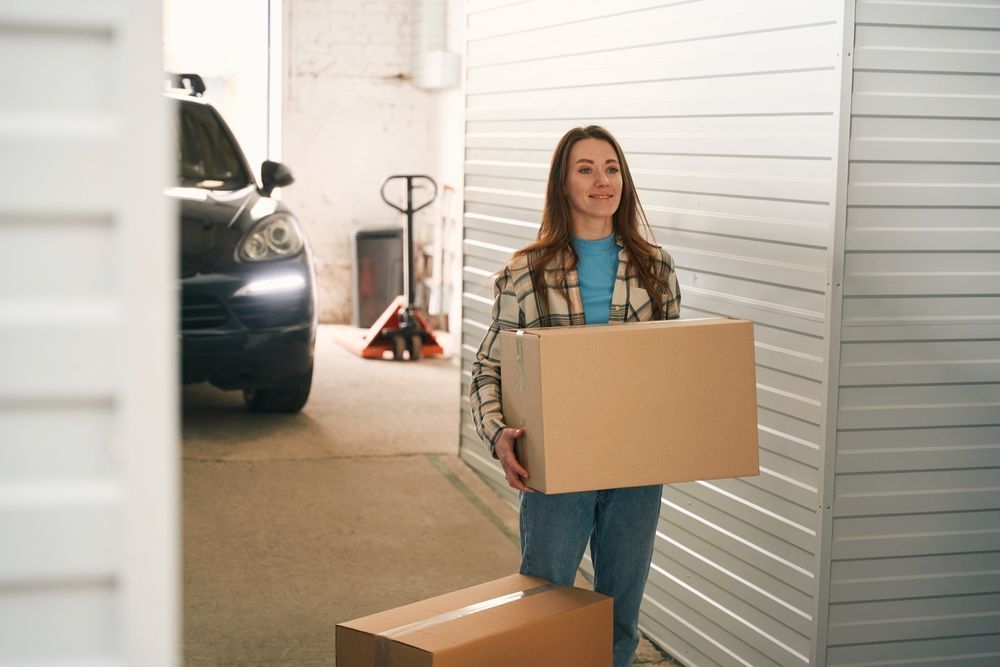 A woman is carrying a cardboard box in a garage.