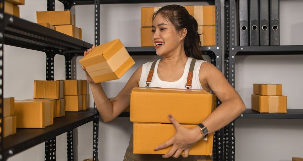 Woman stacking cardboard boxes in a warehouse, holding a box and smiling while organizing shelves
