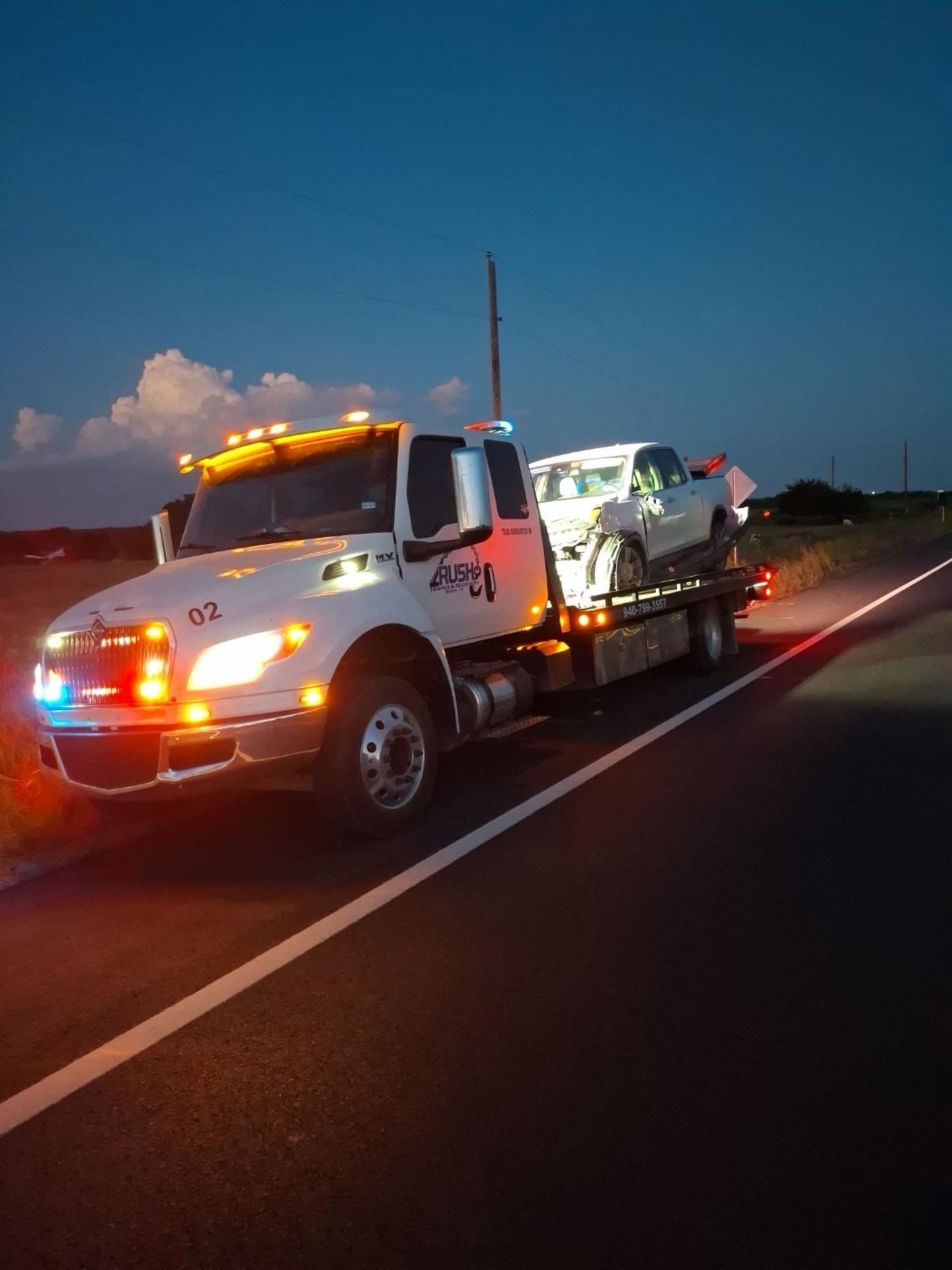 Tow truck hauling a car on a highway at dusk, headlights and hazard lights on