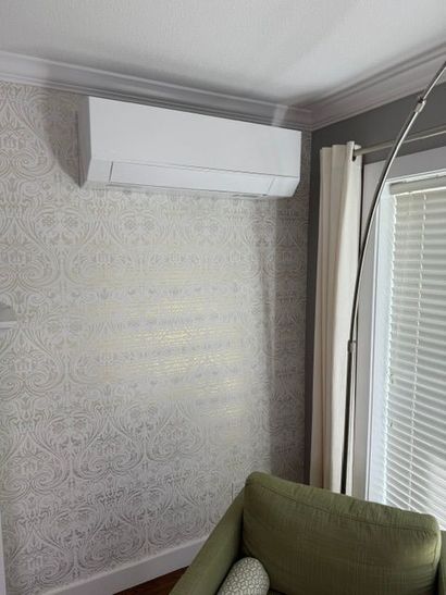 Wall-mounted white air conditioner above patterned wallpaper in a room corner with a green chair and window blinds.