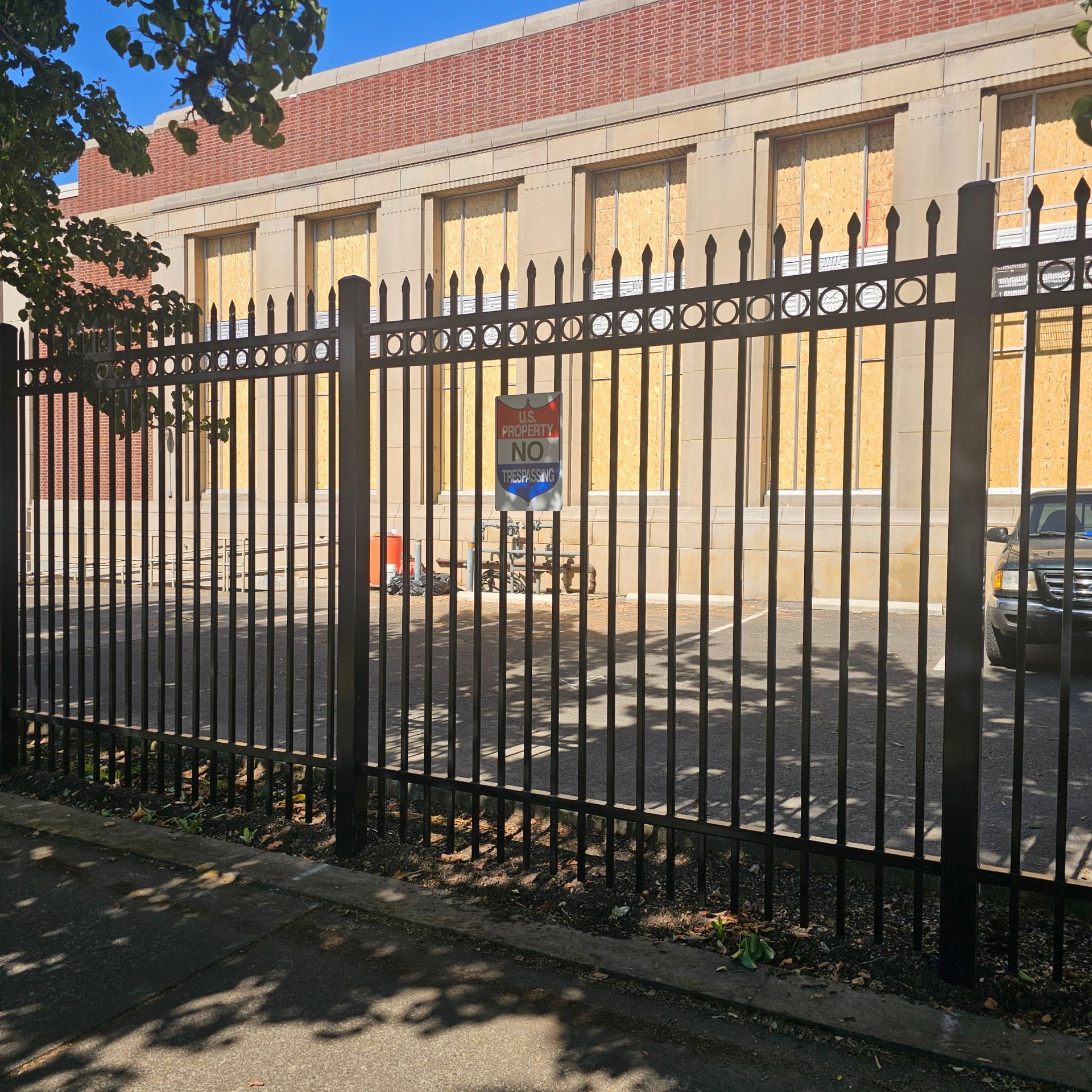 Black metal fence in front of a beige building with boarded windows and a red roof, in sunlight.