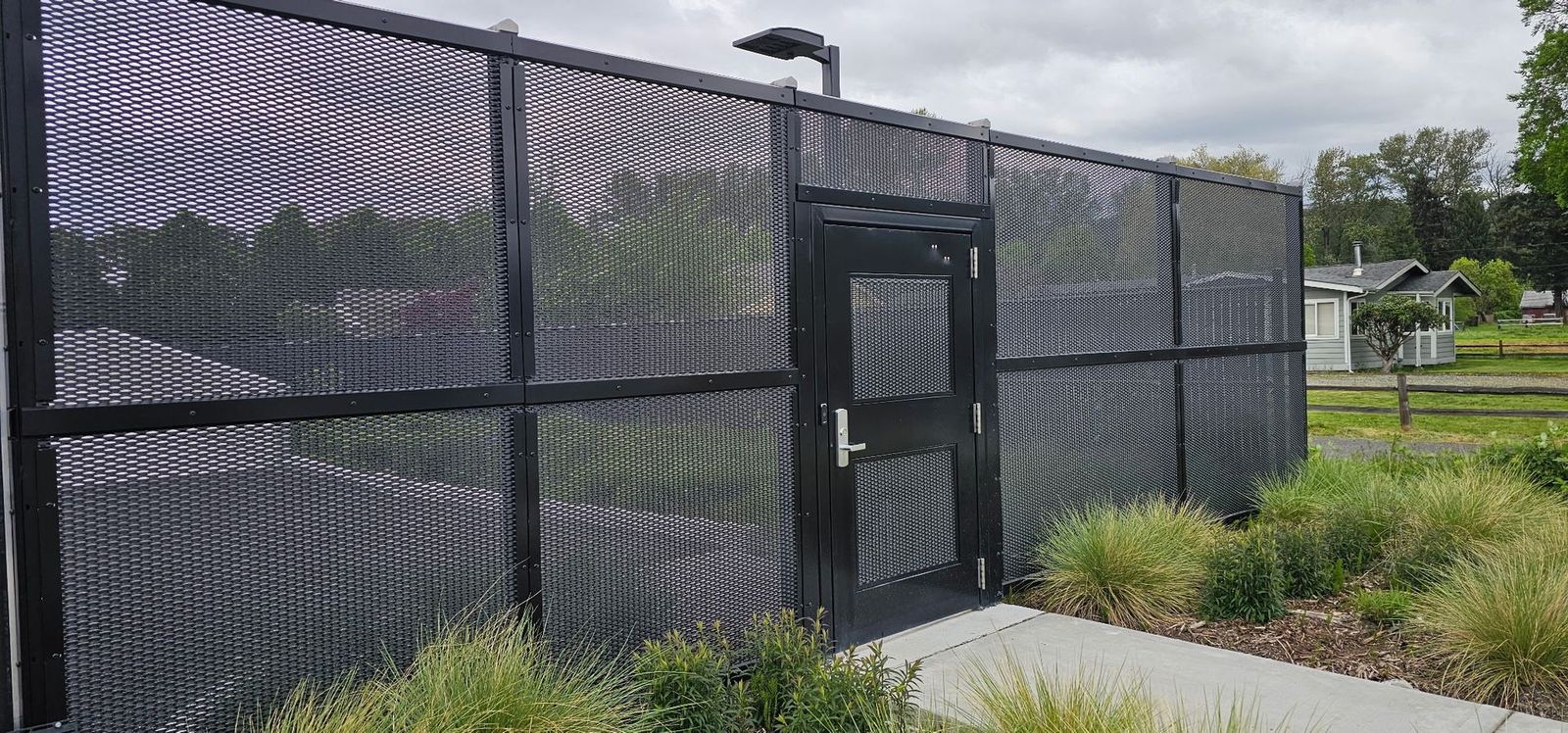 Black chain-link fence with a gate beside a sidewalk and grassy area under cloudy skies