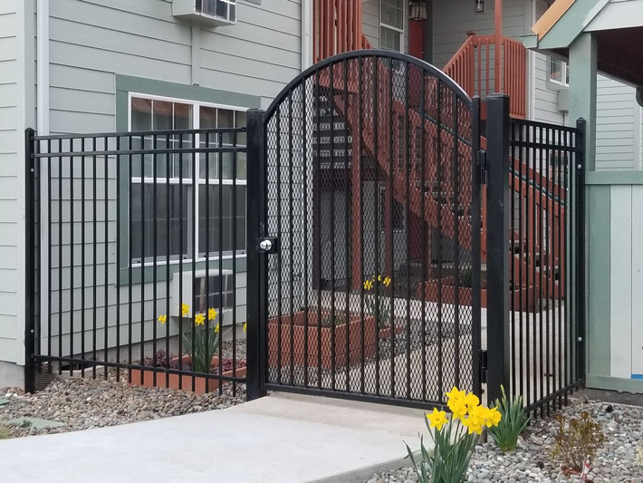 Black metal garden gate and fence in front of a house with brick steps and yellow flowers