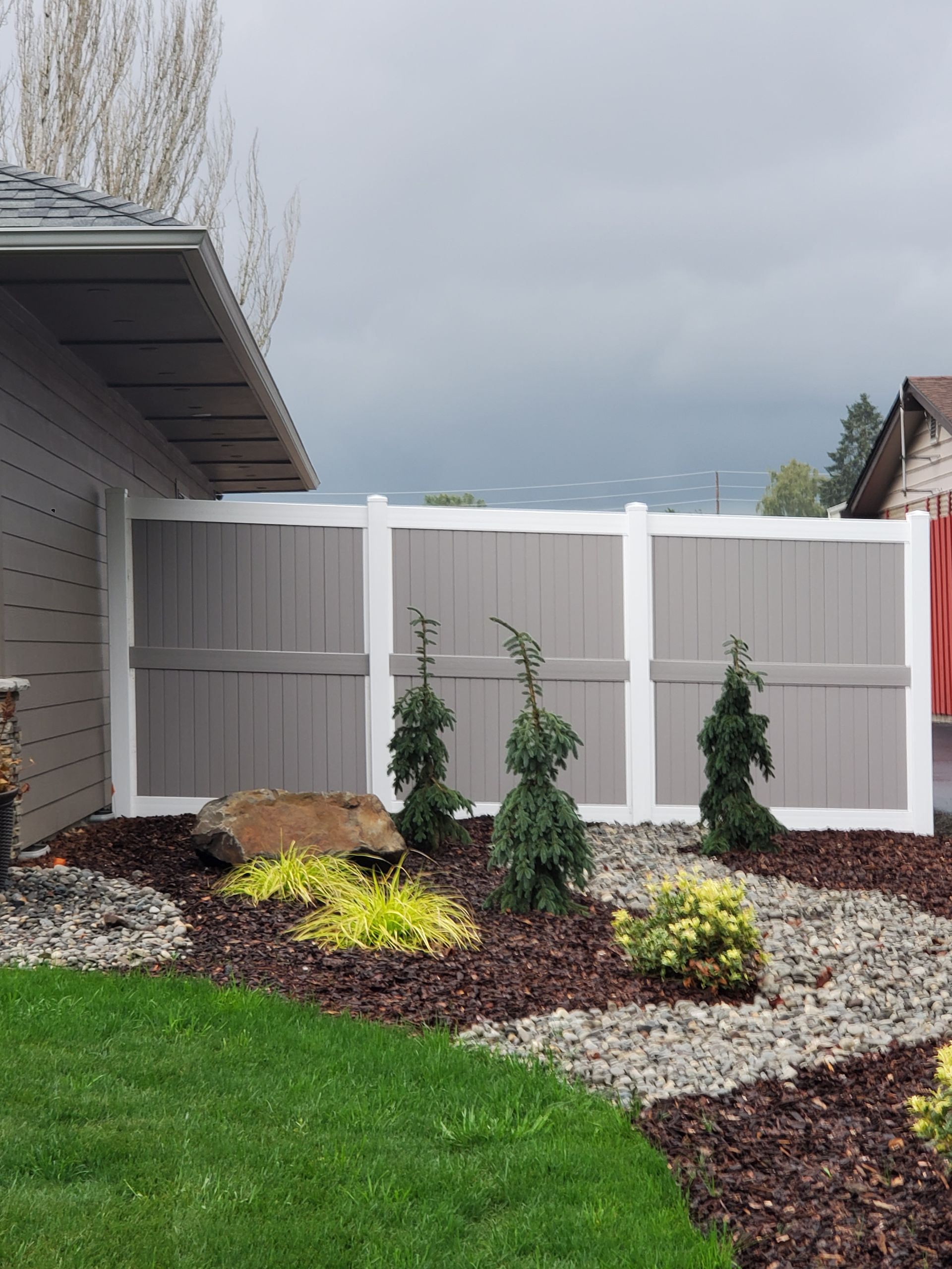 White privacy fence beside a house with new landscaping, shrubs, rocks, mulch, and green lawn