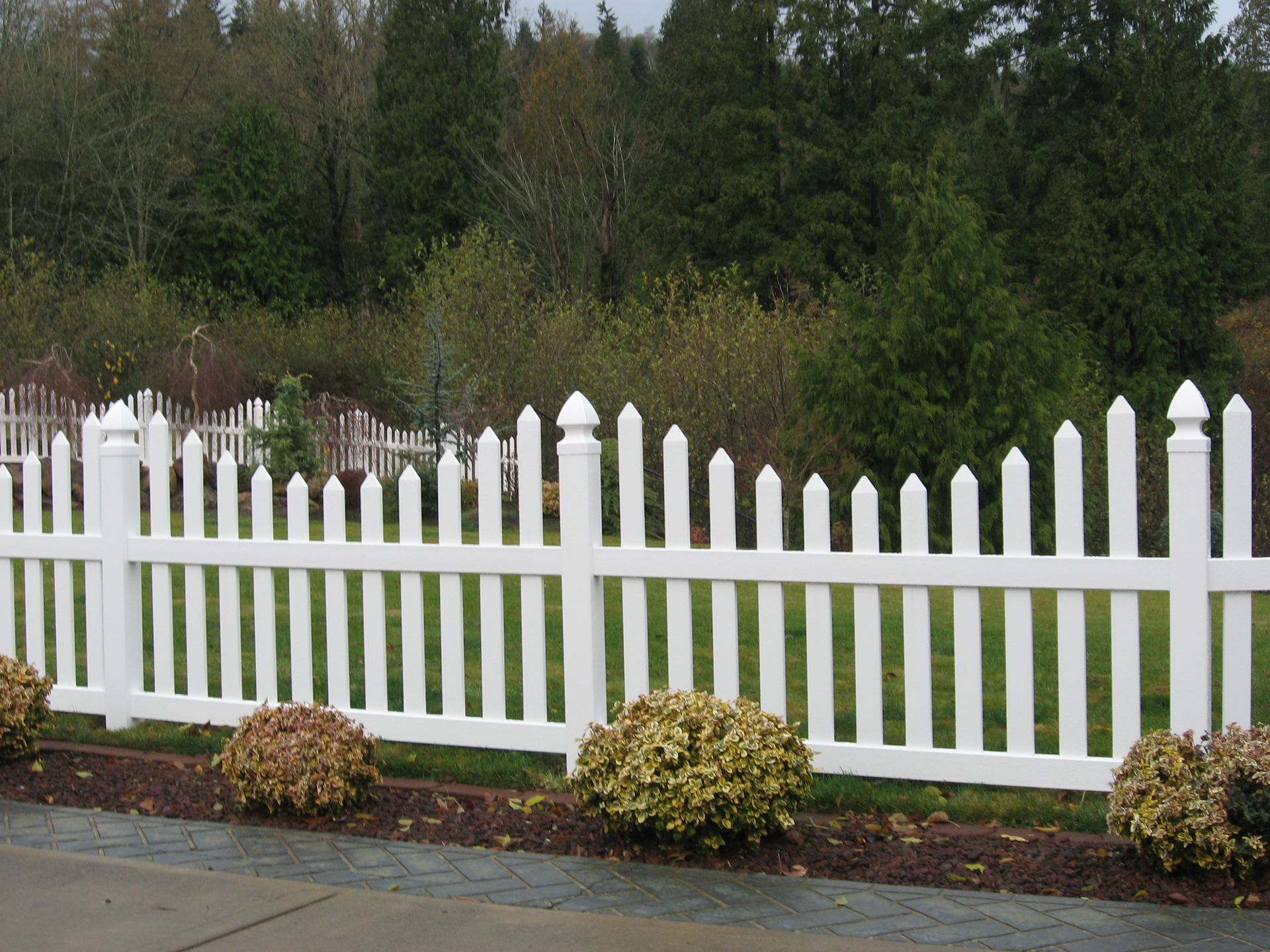 White picket fence with pointed posts along a garden border, backed by trees and shrubs.