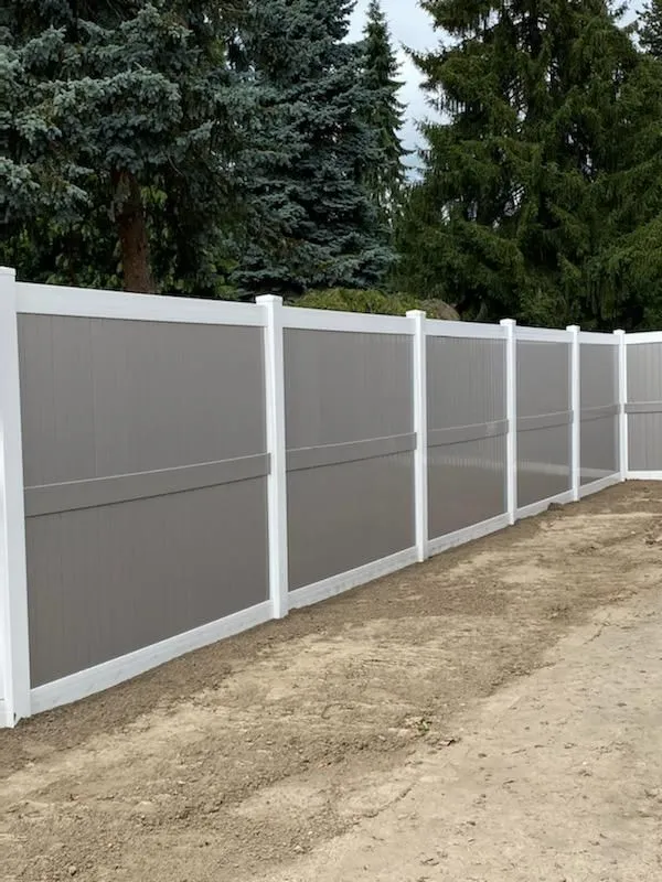 White vinyl fence along a dirt driveway with evergreen trees behind it