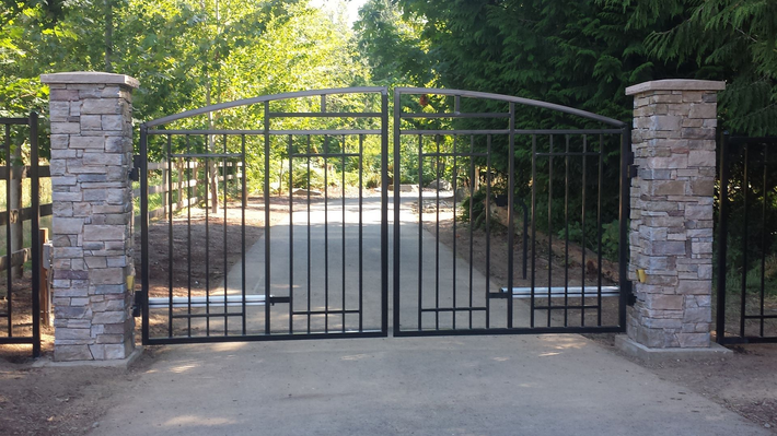 Black metal driveway gate between two stone pillars, with trees and a paved road behind it.