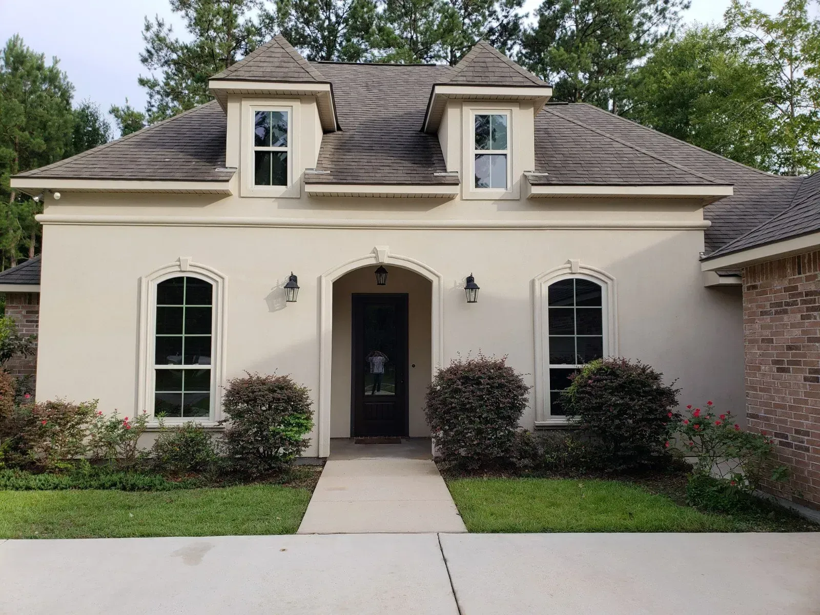 A one-story tan house with a brick accent, two dormer windows, and a central front door surrounded by bushes and a lawn.