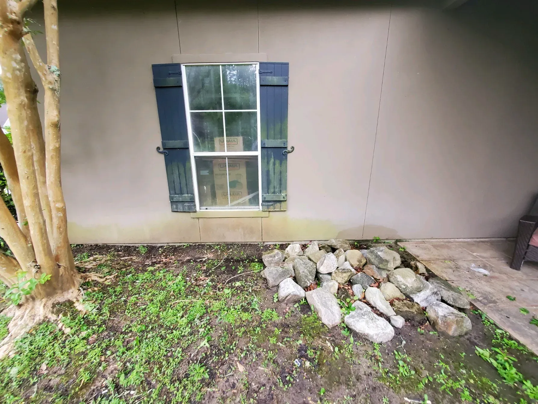 A light-colored stucco exterior wall with a single window featuring dark blue shutters, a tree, and a pile of rocks.