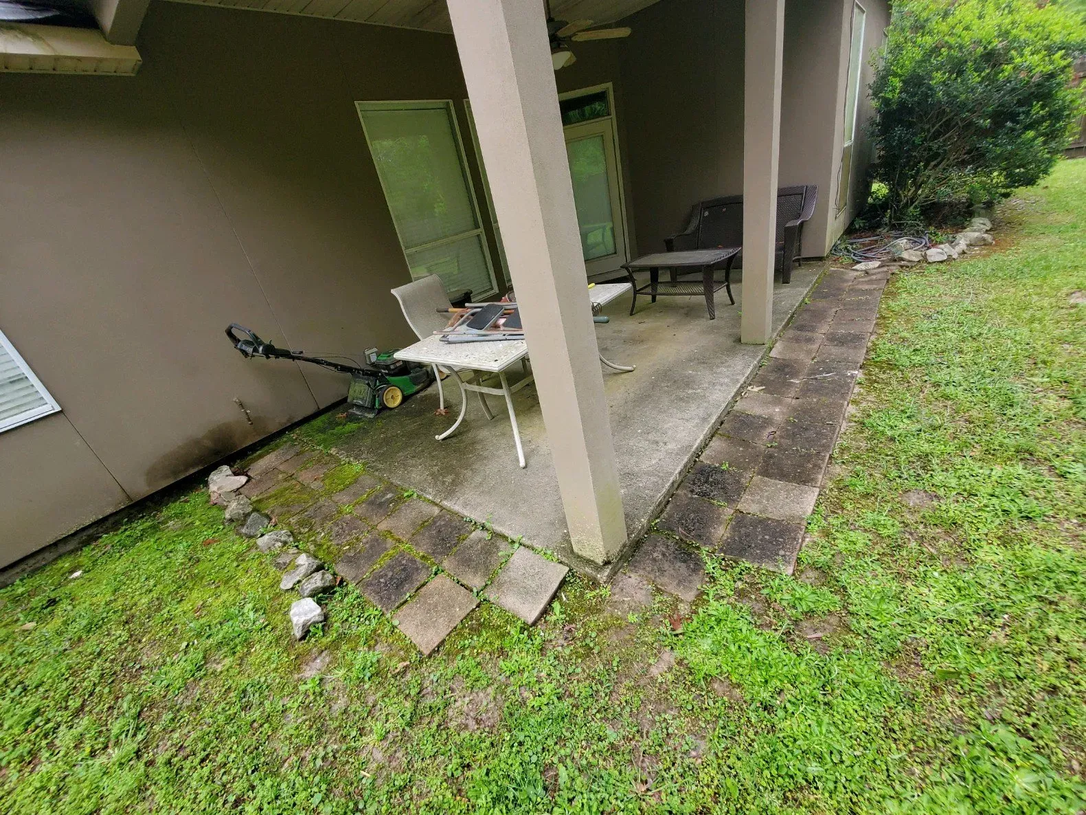 A concrete patio under an overhang features a table, two chairs, and a trimmer on a lawn with stone bordering.