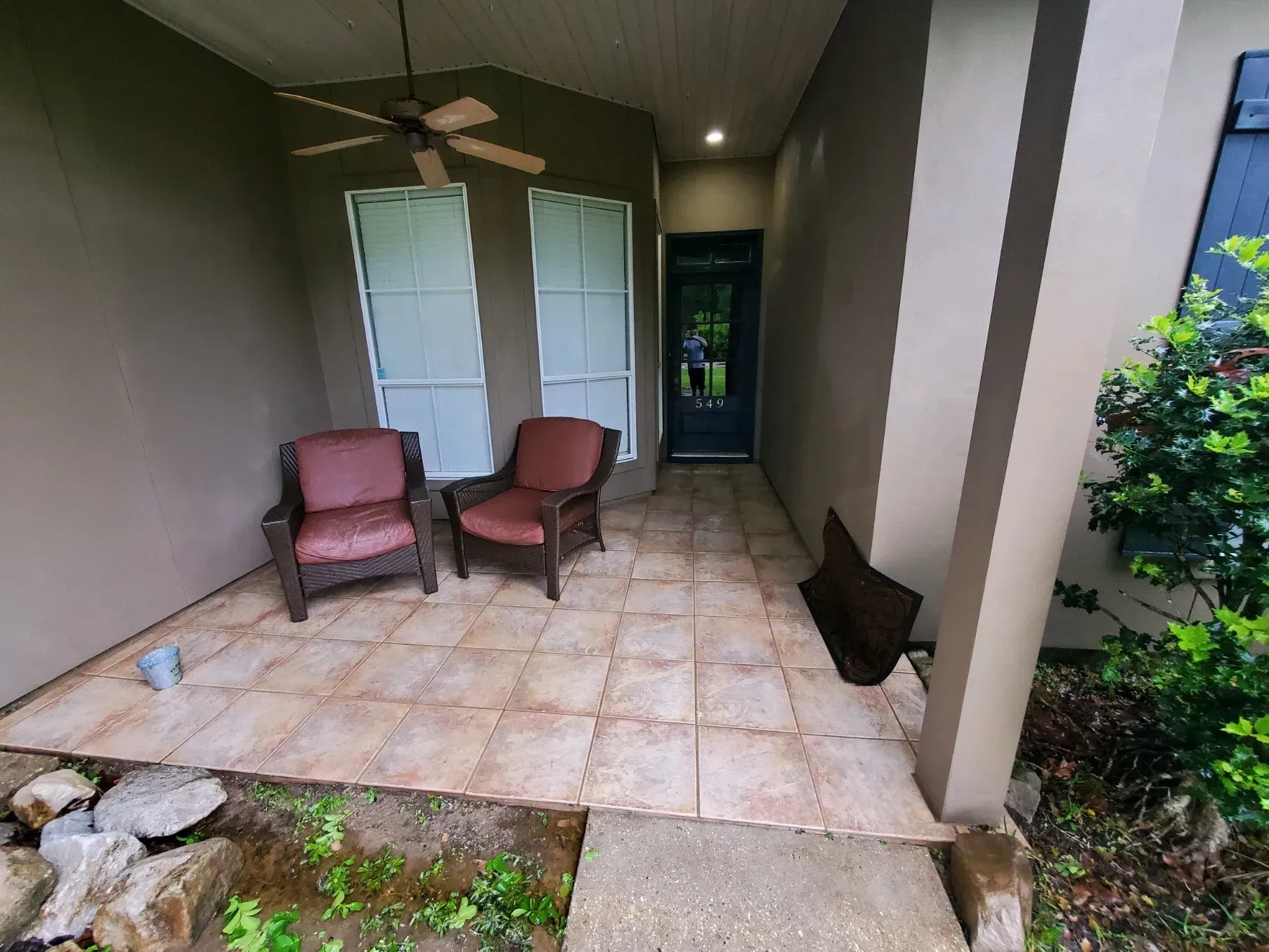 A front porch with two cushioned chairs, a ceiling fan, tiled flooring, and a dark entrance door.